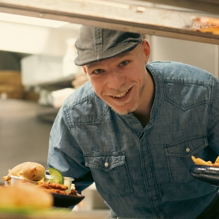 Smiling chef in denim shirt serves freshly cooked meals in a restaurant kitchen.